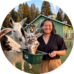 Tutor Sophia standing on a farm, holding a small grain bucket from which three alpacas are eating.